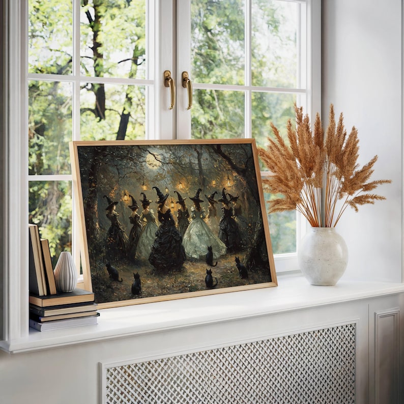 May include: A framed print of a gathering of witches in a moonlit forest. The artwork features figures in long gowns and pointed hats, with black cats scattered around. The print is displayed on a white windowsill with a vase of dried plants.