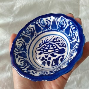 Puede incluir: Un pequeño cuenco de cerámica pintado a mano con un diseño floral azul y blanco. El cuenco tiene un borde festoneado y presenta un motivo de tortuga en el centro. El cuenco se sostiene en una mano.