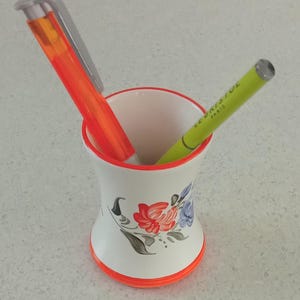 Puede incluir: Un portalápices de cerámica blanca con ribete rojo y naranja, decorado con diseños florales. Un resaltador naranja y un bolígrafo verde con el texto "LE BRISTOL PARIS" están dentro.