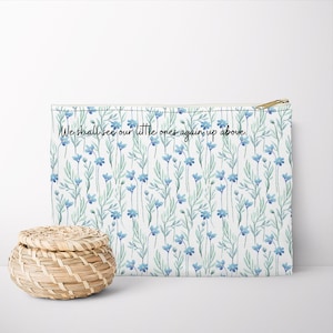 May include: A white zippered pouch with a blue floral pattern and the text "We shall see our little ones again up above." A small, round, woven basket is in front of the pouch.
