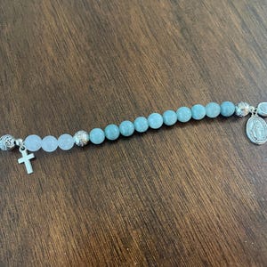 May include: A beaded bracelet with light blue and pink beads, and silver-toned details. The bracelet has a small cross charm and a religious medallion. The clasp is a silver toggle.