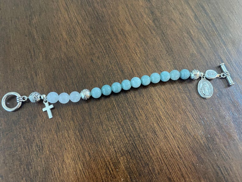May include: A beaded bracelet with light blue and pink beads, and silver-toned details. The bracelet has a small cross charm and a religious medallion. The clasp is a silver toggle.