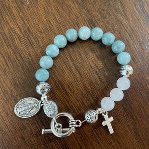 May include: A bracelet with light blue and white beads, featuring silver-toned decorative beads, a cross charm, and two oval pendants. The bracelet is secured with a toggle clasp. The beads are likely made of stone.