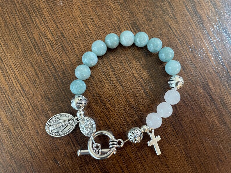 May include: A bracelet with light blue and white beads, featuring silver-toned decorative beads, a cross charm, and two oval pendants. The bracelet is secured with a toggle clasp. The beads are likely made of stone.