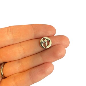 May include: A small, silver-coloured charm with a circular design. The charm features two baby footprints inside the circle. The charm is held in a hand against a white background.