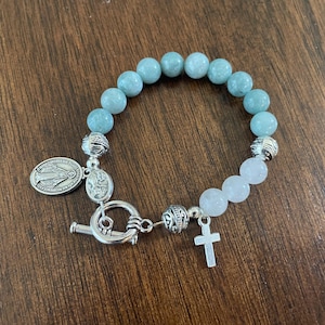 May include: A beaded bracelet with light blue and white round stones. The bracelet has a silver-tone toggle clasp, a small cross charm, and a religious medal. The bracelet is displayed on a wooden surface.