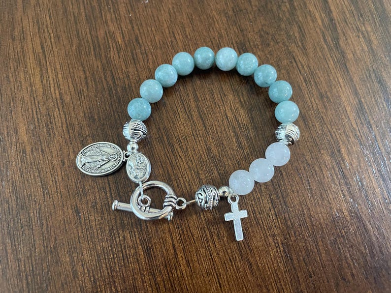 May include: A beaded bracelet with light blue and white round stones. The bracelet has a silver-tone toggle clasp, a small cross charm, and a religious medal. The bracelet is displayed on a wooden surface.