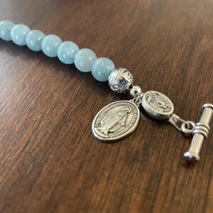 May include: A light blue beaded bracelet with silver-toned details. The bracelet has round, light blue beads and a silver clasp with two oval pendants and a toggle closure. The pendants feature religious imagery.