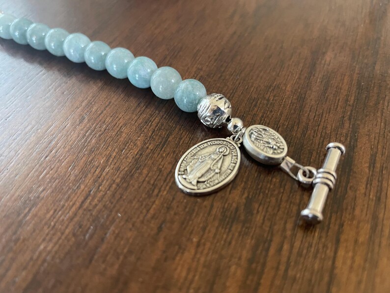 May include: A light blue beaded bracelet with silver-toned details. The bracelet has round, light blue beads and a silver clasp with two oval pendants and a toggle closure. The pendants feature religious imagery.