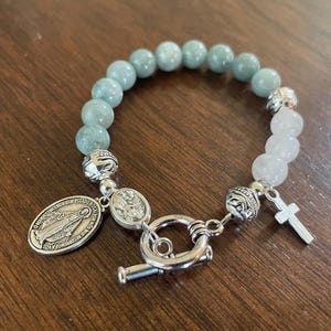 May include: A bracelet with light blue and white beads, featuring a silver cross charm and an oval pendant with an image of the Virgin Mary. The bracelet is secured with a silver toggle clasp. The beads are approximately 0.8 cm in diameter.