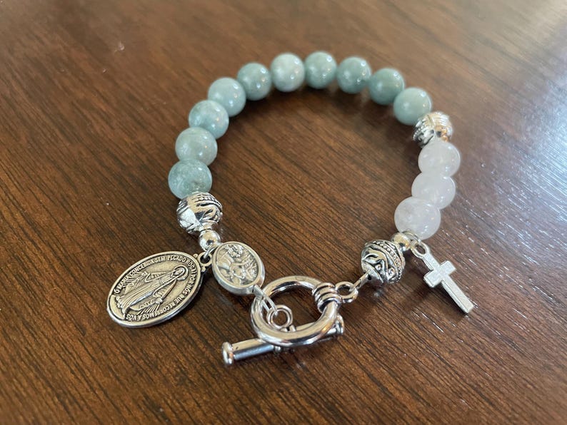 May include: A bracelet with light blue and white beads, featuring a silver cross charm and an oval pendant with an image of the Virgin Mary. The bracelet is secured with a silver toggle clasp. The beads are approximately 0.8 cm in diameter.