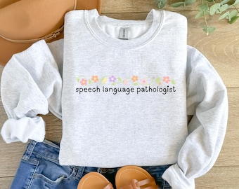 May include: A pale grey sweatshirt featuring the words "speech language pathologist" in black, with a pastel floral design above. The sweatshirt is laid on blue jeans, a brown hat, and sandals.