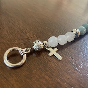 May include: A beaded bracelet with a silver cross charm and a key ring clasp. The bracelet features grey and white beads, with decorative silver beads separating the sections. The background is a dark wood surface.