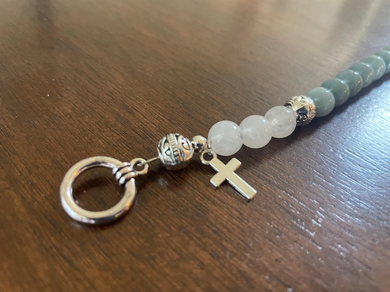 May include: A beaded bracelet with a silver cross charm and a key ring clasp. The bracelet features grey and white beads, with decorative silver beads separating the sections. The background is a dark wood surface.