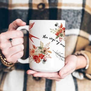 Op de afbeelding: Witte keramische mok met bloemen- en veermotieven en de tekst "Then sings my soul". De mok heeft een groot handvat en is versierd met bloemen in aquarel-stijl in tinten rood, oranje en bruin. Een rode vogel is ook afgebeeld op de mok.