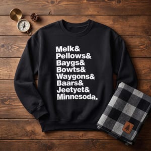Puede incluir: Una sudadera negra de manga larga con cuello redondo y el texto blanco Melk& Pellows& Baygs& Bowts& Waygons& Baars& Jeetyet& Minnesoda. La prenda se muestra junto a una brújula y una manta de cuadros negros y blancos.