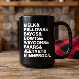Puede incluir: Una taza de cerámica negra mate con texto blanco que dice: MELK& PELLOWS& BAYGS& BOWTS& WAYGONS& BAARS& JEETYET& MINNESODA. La taza tiene un asa estándar y un cuerpo cilíndrico, mostrando una lista de palabras en una fuente sans-serif en negrita.