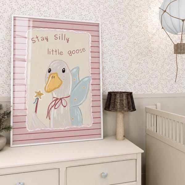 Puede incluir: Una lámina enmarcada que muestra una oca blanca con pico amarillo, alas de hada azules y una varita con una estrella. El texto "stay silly little goose" aparece sobre un fondo crema con un borde de rayas rosas.
