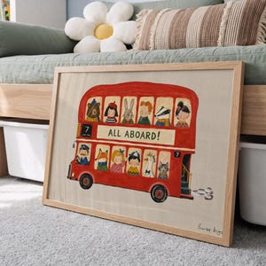 May include: A framed print featuring a red double-decker bus illustration with the text ALL ABOARD! and the number 7. The bus is filled with various illustrated animal and human characters looking out of the windows, set against a light background.