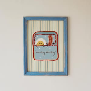 May include: A framed print featuring a cartoon illustration of a fried egg and a strip of bacon tucked into a light blue bed with the text Wakey Wakey! The artwork is set against a cream background with thin vertical blue stripes and a blue frame.