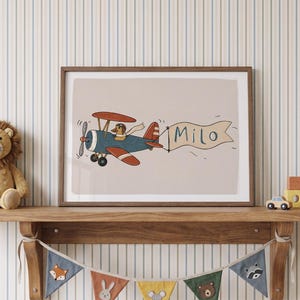 May include: A framed horizontal print featuring a blue and red biplane piloted by a duck wearing a pilot cap and scarf. The plane pulls a banner with the name Milo written in blue. The illustration is set against a light beige background.