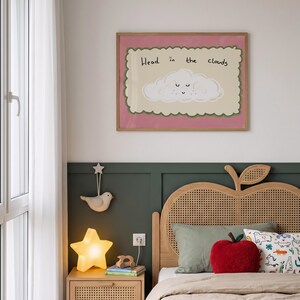May include: A framed wall art print featuring a smiling cloud illustration and the text Head in the clouds, displayed above a wooden bed with a cane headboard, a red apple-shaped pillow, a star-shaped lamp, and a hanging bird ornament.