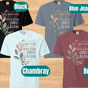 May include: Four t-shirts in different colours: black, blue jean, chambray, and brick. Each shirt features a watercolour-style floral design with the text "Saved By Grace Alone Through Faith Alone In Christ Alone Ephesians 2:8".