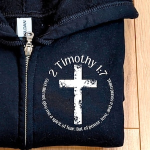 Puede incluir: Sudadera con capucha negra con cremallera y un gr&aacute;fico circular blanco con una cruz y el texto "2 Timoteo 1:7 Dios no me dio un esp&iacute;ritu de miedo, sino un esp&iacute;ritu de poder, amor y una mente sana."