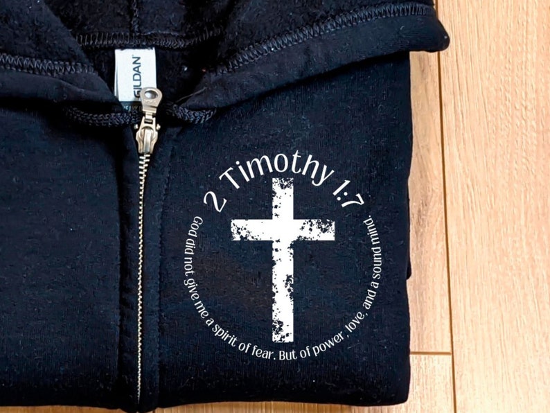 Puede incluir: Sudadera con capucha negra con cremallera y un gr&aacute;fico circular blanco con una cruz y el texto "2 Timoteo 1:7 Dios no me dio un esp&iacute;ritu de miedo, sino un esp&iacute;ritu de poder, amor y una mente sana."