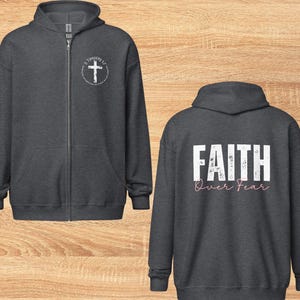 Puede incluir: Dos sudaderas con capucha de color gris oscuro con cremallera. Una sudadera tiene una cruz blanca con el texto "2 Timoteo 1:7" en un c&iacute;rculo en el pecho izquierdo. La otra sudadera tiene el texto "FAITH over FEAR" en blanco con letras rosas en la espalda.
