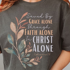 May include: A dark grey t-shirt with a floral design and the text "Saved By Grace Alone Through Faith Alone In Christ Alone Ephesians 2:8".