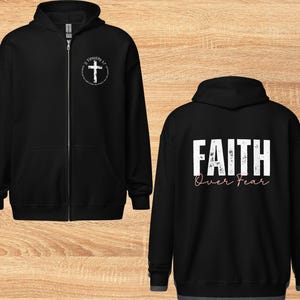 Puede incluir: Sudadera con capucha negra con cremallera, con una cruz blanca y el texto "2 Timoteo 1:7" en la parte delantera y el texto "FAITH Over Fear" en la parte trasera.