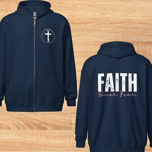 Puede incluir: Dos sudaderas con capucha azul marino con cremallera. La sudadera delantera tiene una cruz blanca con el texto "2 Timoteo 1:7" en un c&iacute;rculo. La sudadera trasera tiene el texto "FAITH over FEAR" en blanco con letras rosas.