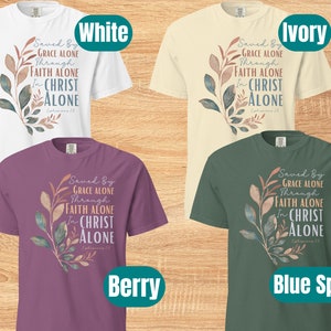 May include: Four t-shirts in different colours with a floral design and the text "Saved By Grace Alone Through Faith Alone In Christ Alone Ephesians 2:8". The colours are white, ivory, berry, and blue spruce.