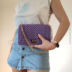 Può includere: Borsa a mano all'uncinetto viola fatta a mano con catena e chiusura dorate. La borsa presenta un motivo a spina di pesce e una forma rettangolare. Il sito web ainaoracrochet.com è visibile.