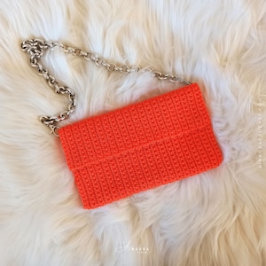 Puede incluir: Bolso de mano de ganchillo naranja brillante con una cadena plateada. El bolso rectangular está hecho de un material tejido texturizado. El bolso de mano está sobre un fondo blanco y esponjoso.
