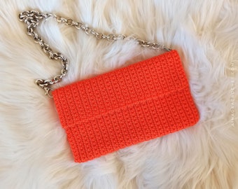 Könnte beinhalten: Hellorangefarbene Häkeltasche mit silberner Kette. Die rechteckige Tasche besteht aus einem strukturierten, gestrickten Material. Die Clutch liegt auf einem flauschigen weißen Hintergrund.