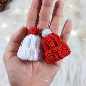 May include: Two miniature crocheted hats, one white and one red, each with a fluffy pom-pom. The hats are held in a hand, showcasing the intricate stitch work and festive design. The red hat has a red pom-pom, and the white hat has a white pom-pom.