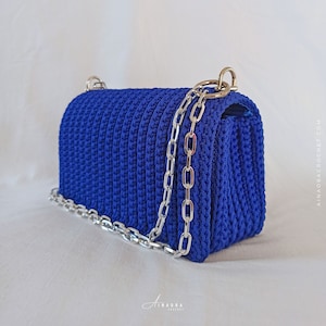 May include: A cobalt blue crochet handbag with a silver chain strap. The bag has a rectangular shape with rounded edges and a textured surface. The silver chain is attached to the bag with silver rings. The bag is on a white background.
