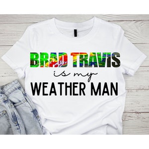 Puede incluir: Camiseta blanca con el texto "Brad Travis is my Weather Man" en negro. La palabra "Brad Travis" está en una fuente colorida similar a un radar meteorológico.