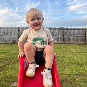 May include: A young boy wearing a beige t-shirt with a green caterpillar graphic and the text "Still Hungry" sits on a red plastic slide. He is smiling and wearing white sneakers with blue and yellow accents.