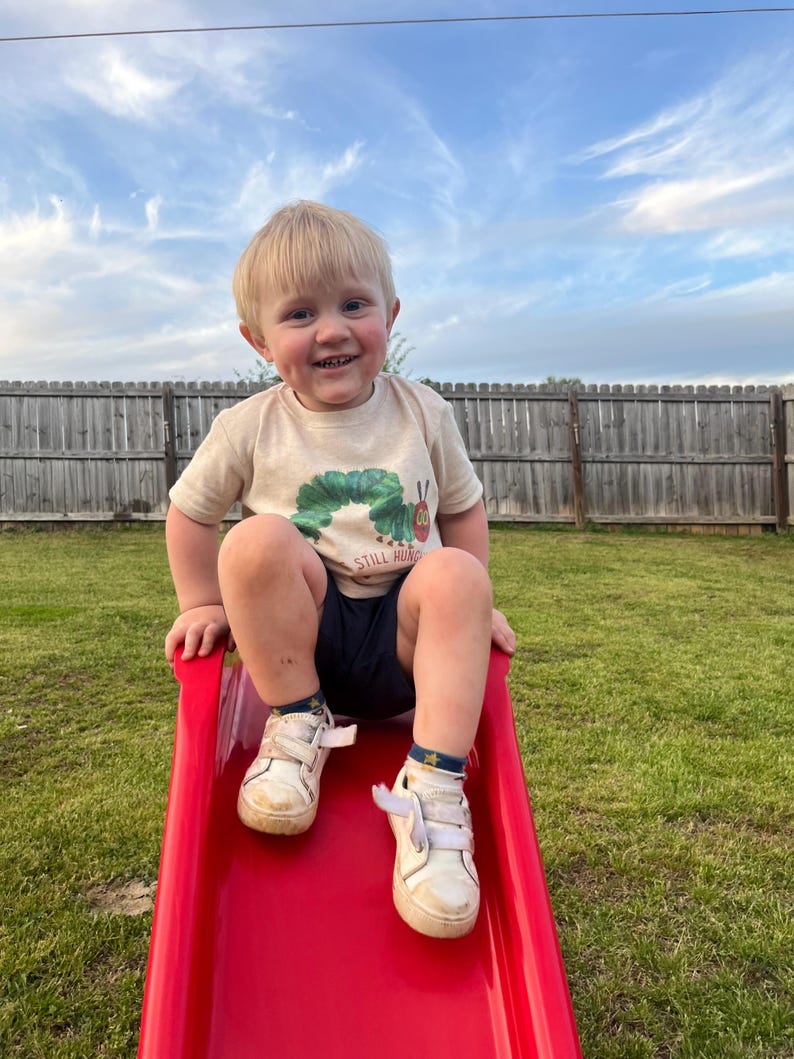 May include: A young boy wearing a beige t-shirt with a green caterpillar graphic and the text "Still Hungry" sits on a red plastic slide. He is smiling and wearing white sneakers with blue and yellow accents.