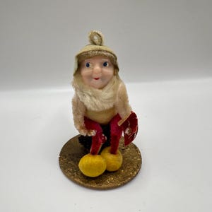 May include: A vintage figurine of a seated gnome with a painted face, wearing a beige hat, scarf, and fur-like body. The gnome has red pants, yellow feet, and holds a red shield. It sits on a gold-colored, round base.