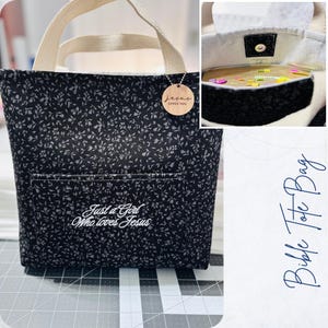 May include: A black floral print tote bag with cream handles and a front pocket. The bag features the embroidered text "Just a Girl Who loves Jesus". A wooden tag with "Jesus loves you" hangs from the handle. The bag is designed for carrying a Bible.