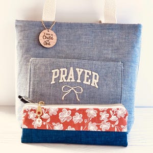 May include: A blue tote bag with a cream-colored handle and a front pocket embroidered with the word "PRAYER". A wooden tag with the text "I am a Child of God" hangs from the handle. A floral pencil case with a gold zipper is in front of the bag.