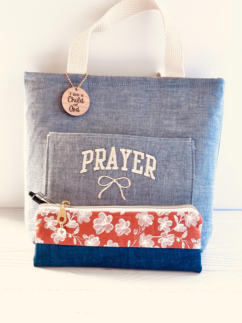 May include: A blue tote bag with a cream-colored handle and a front pocket embroidered with the word "PRAYER". A wooden tag with the text "I am a Child of God" hangs from the handle. A floral pencil case with a gold zipper is in front of the bag.