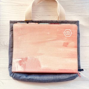 May include: A pink and peach-colored journal with a watercolor design rests inside a gray tote bag with a cream-colored handle. The journal has a white logo in the corner. The tote bag is made of a denim-like material.