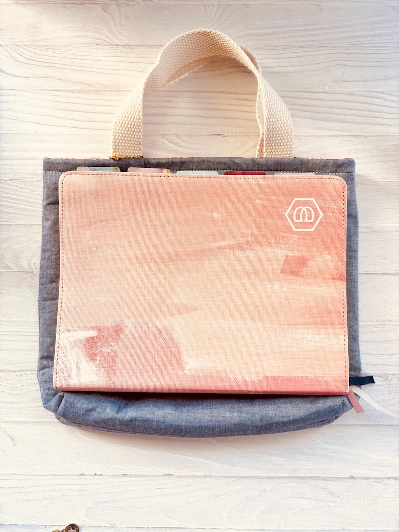 May include: A pink and peach-colored journal with a watercolor design rests inside a gray tote bag with a cream-colored handle. The journal has a white logo in the corner. The tote bag is made of a denim-like material.