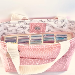 May include: Pink tote bag with white polka dots and cream-coloured handles. The interior lining features a butterfly pattern. The bag is open, revealing a tray filled with small, colourful cards.