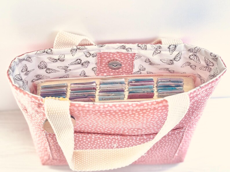 May include: Pink tote bag with white polka dots and cream-coloured handles. The interior lining features a butterfly pattern. The bag is open, revealing a tray filled with small, colourful cards.